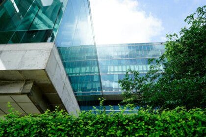 Bâtiment moderne en verre et en béton, entouré de verdure avec un ciel bleu partiellement nuageux en arrière-plan.