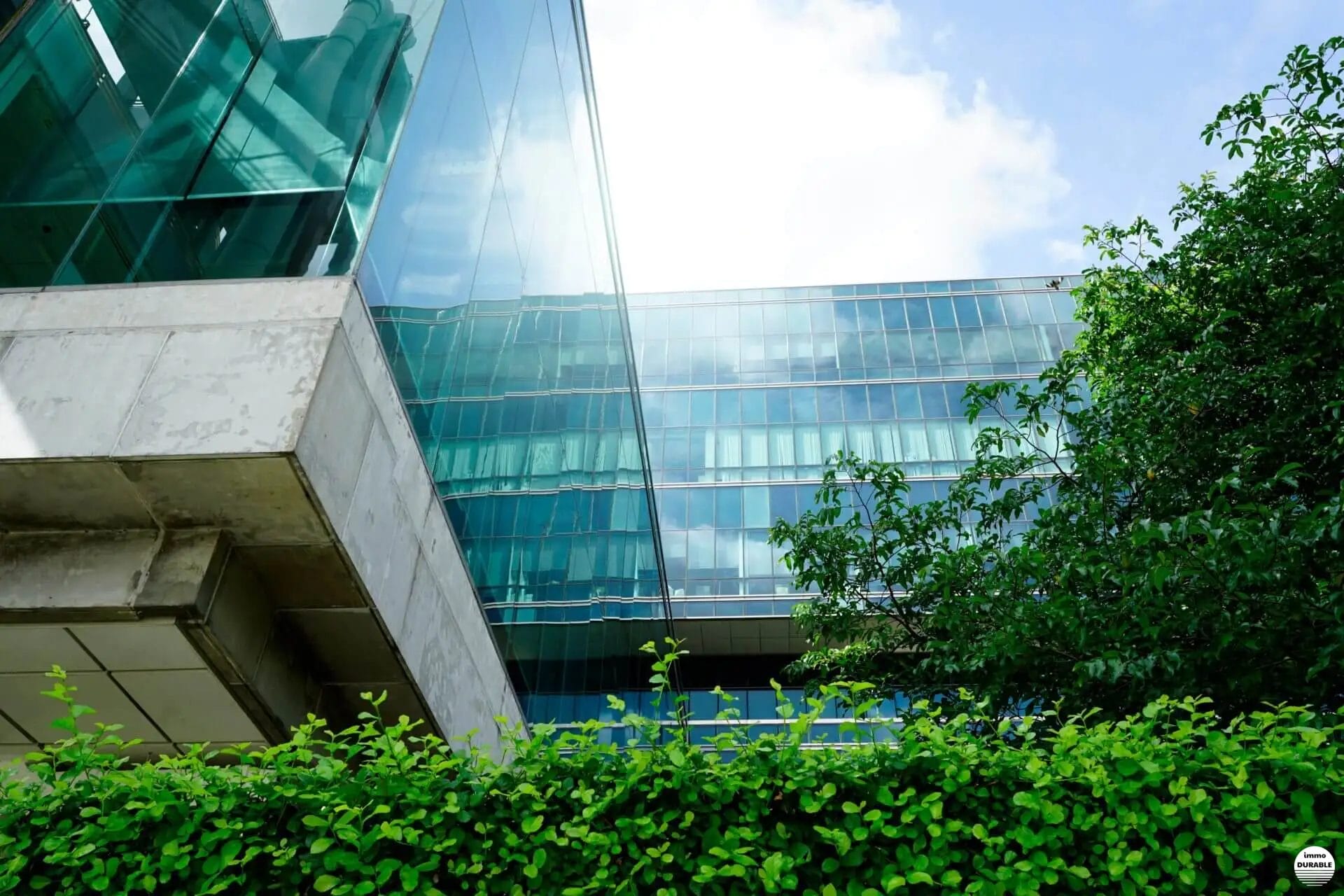 Bâtiment moderne en verre et en béton, entouré de verdure avec un ciel bleu partiellement nuageux en arrière-plan.